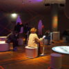 Ausstellungsbereich Leipzig 89 im Deutschen Historischen Museum Berlin. In der Mitte eine runde Projektionsfläche, darum kreisförig angeordnet Objekte, Tablets und Sitzflächen. Einige Besucher*innen sitzen und spielen auf mobilen Geräten, andere stehen und unterhalten sich