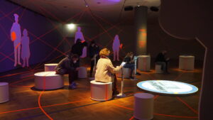Ausstellungsbereich Leipzig 89 im Deutschen Historischen Museum Berlin. In der Mitte eine runde Projektionsfläche, darum kreisförig angeordnet Objekte, Tablets und Sitzflächen. Einige Besucher*innen sitzen und spielen auf mobilen Geräten, andere stehen und unterhalten sich