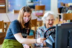 Alte Frau spielt mit Lenkrad ein digitales Autorennspiel. Foto Autorennen Lenkrad: Alpha STORYTELLING by Kai Kapitän