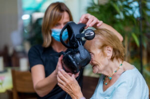 Gaming im Altenzentrum: Foto einer alten Frau, der gerade eine Virtual Reality Brill aufgesetzt wird.