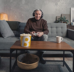 Titelbild zum PlayingHistory Blogbeitrag "Gaming im Altenzentrum - Mut zur digitalen Teilhabe". Alter Mann auf Couch mit Controller in der Hand.